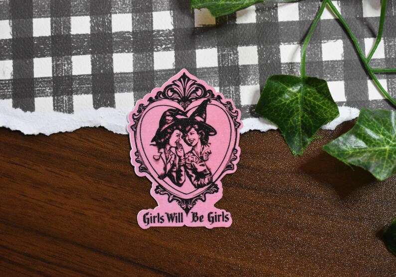 May include: A pink sticker featuring a black illustration of two women in witch hats within a heart-shaped frame. The text "Girls Will Be Girls" is at the bottom. The sticker is on a wooden surface with a checkered pattern in the background.