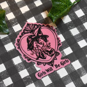 May include: A pink sticker with a heart-shaped design featuring two witches in black and white. The sticker has the text "Girls Will Be Girls" in a pink banner. The background is a black and white checkered pattern.