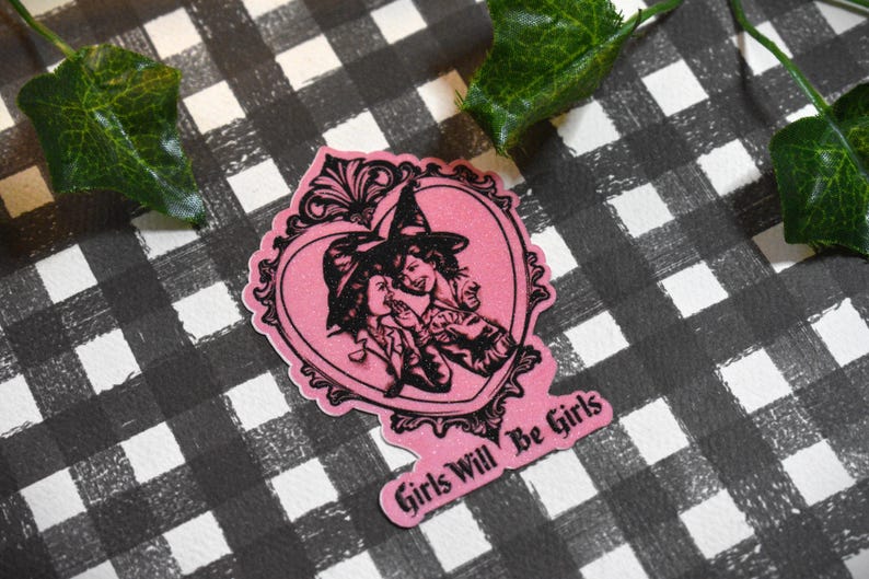 May include: A pink sticker with a heart-shaped design featuring two witches in black and white. The sticker has the text "Girls Will Be Girls" in a pink banner. The background is a black and white checkered pattern.