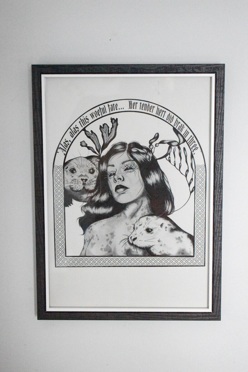 May include: Black and white framed art print featuring a woman with long hair and two seals. The artwork includes text in an arch above the woman. The frame is dark brown.