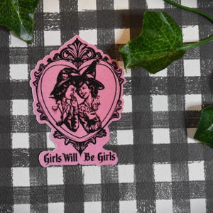 May include: A pink sticker featuring a black illustration of two women in witch hats within a heart-shaped frame. The text "Girls Will Be Girls" is below. Set against a black and white checked pattern with green leaves.