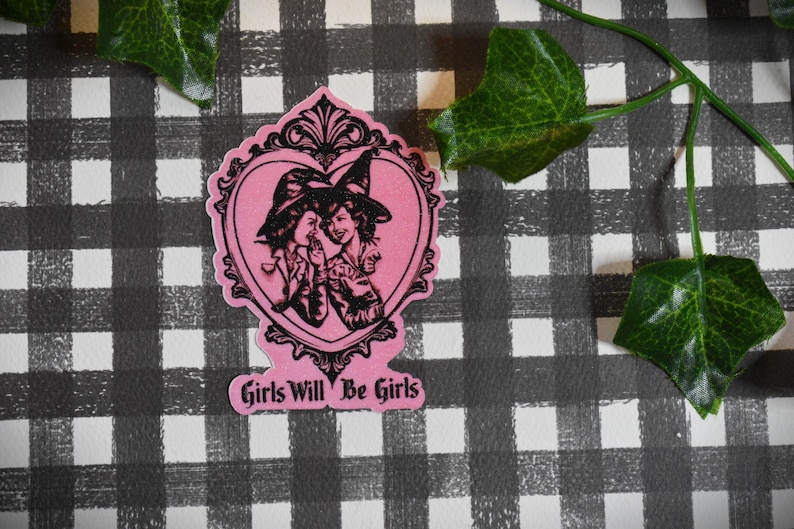 May include: A pink sticker featuring a black illustration of two women in witch hats within a heart-shaped frame. The text "Girls Will Be Girls" is below. Set against a black and white checked pattern with green leaves.