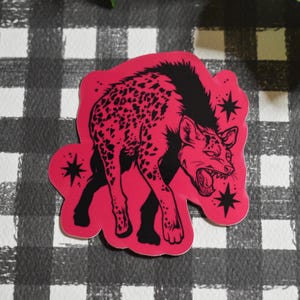 May include: A die-cut sticker featuring a vibrant hot pink hyena with black spots and a black mane. The hyena is depicted in a snarling pose with its mouth open, surrounded by small black star-shaped accents on a bright pink background.