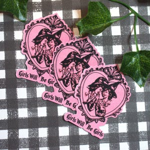 May include: Pink stickers with a heart-shaped frame featuring two women in witch hats whispering. The stickers have the text "Girls Will Be Girls" in a cursive font. The stickers are on a black and white checkered surface.