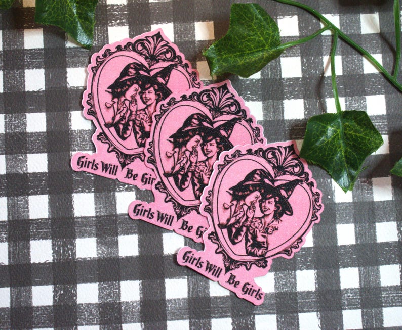 May include: Pink stickers with a heart-shaped frame featuring two women in witch hats whispering. The stickers have the text "Girls Will Be Girls" in a cursive font. The stickers are on a black and white checkered surface.