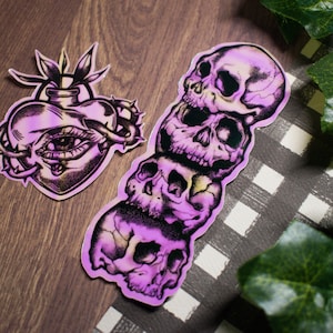 May include: Two stickers with a gothic theme. One sticker features a heart with an eye inside, surrounded by thorns. The other sticker features a stack of three skulls.