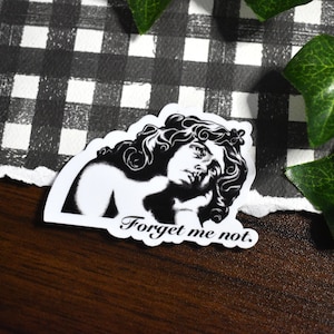May include: A black and white sticker featuring a classical bust with the text "Forget me not." The sticker has a white border and is placed on a dark wooden surface with a black and white checkered pattern in the background.