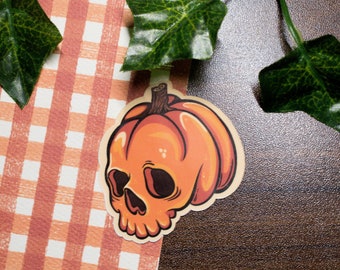 May include: A sticker of an orange pumpkin with a skull face. The sticker is on a checkered background.
