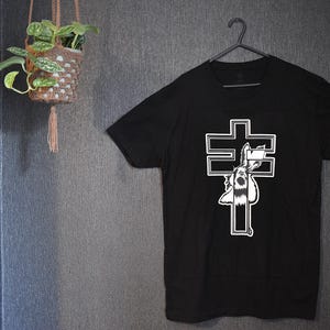 Puede incluir: Camiseta negra con un gráfico blanco de un conejo dentro de un diseño de cruz. La camiseta está colgada en una percha negra contra una pared gris oscura. Una planta colgante en una cesta tejida es visible en el fondo.