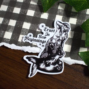 May include: A black and white sticker featuring a mermaid illustration with the text "Trust in New Beginnings." The mermaid is depicted with detailed scales and flowing hair. The sticker is on a checkered background.