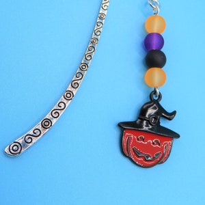 May include: Silver bookmark with a decorative swirl pattern and a chain attached to a black pumpkin with a witch's hat and orange, purple, and black beads.