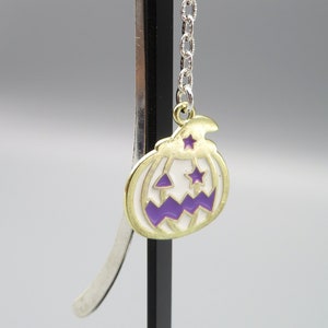 May include: A silver bookmark with a chain and a gold pumpkin charm. The pumpkin is white with a purple face and two stars.