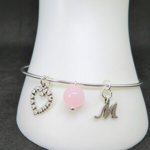 Puede incluir: Un brazalete de plata con tres abalorios: un corazón, una cuenta rosa y una letra M.