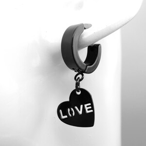 Puede incluir: Pendiente de aro negro con un dije de corazón colgante que dice "LOVE" en letras blancas.