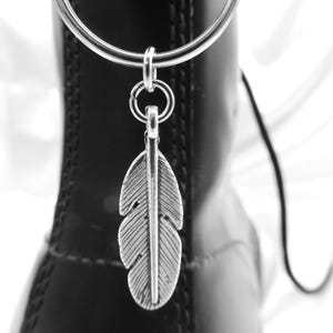 以下が含まれることがあります： 黒いブーツに付いたシルバーのリングから、銀色の羽根のチャームがぶら下がっています。チャームにはエッチングされた線があり、ブーツにはステッチと光沢のある仕上げが見えます。背景は柔らかな白です。
