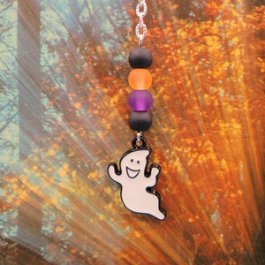May include: A silver bookmark with a white ghost charm and colorful beads. The beads are black, orange, and purple. The ghost has a smiling face.