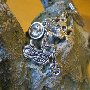 May include: Silver-coloured keychain featuring motorcycle, skull and crossbones, and yin-yang charms. The motorcycle charm has detailed wheels, a seat, and handlebars. The skull and crossbones charm is a classic biker symbol. The yin-yang charm adds balance.
