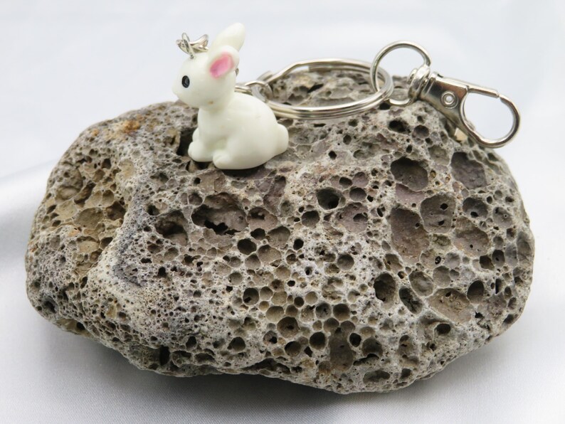 May include: A white rabbit keychain with a pink nose and a silver crown. The keychain is attached to a silver key ring and a silver clasp.