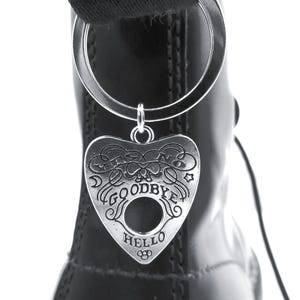 May include: A silver-toned metal heart-shaped planchette keychain attached to a black boot pull tab. The planchette features engraved text including YES, NO, GOODBYE, and HELLO, along with decorative swirls, stars, and a central circular cutout.