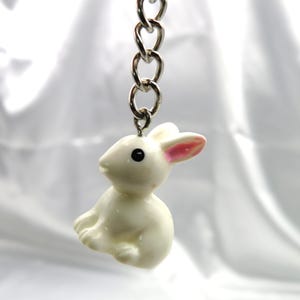 May include: A white rabbit keychain with pink ears and black eyes. The keychain is attached to a silver chain.