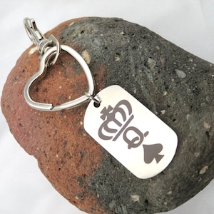 May include: A silver-colored keychain with a heart-shaped ring and a rectangular tag. The tag features a crown, the letter "Q", and a spade symbol. The keychain is resting on a textured, multi-colored rock.