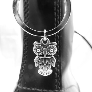 May include: A silver owl charm hangs from a silver ring attached to a black boot. The owl charm is detailed with textured feathers and large eyes. The boot is made of shiny black leather.