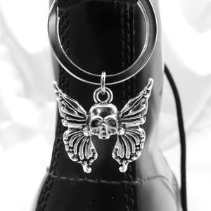 May include: A silver-colored boot charm featuring a skull with butterfly wings, hanging from a metal ring. The charm is attached to a black boot with a shiny finish. The background is white.