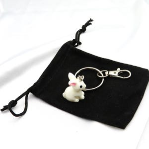 May include: A white ceramic rabbit keychain with pink ears and a black eye. The keychain is attached to a silver key ring and a silver clip. The keychain is sitting on a black velvet pouch with a drawstring.