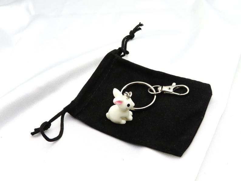 May include: A white ceramic rabbit keychain with pink ears and a black eye. The keychain is attached to a silver key ring and a silver clip. The keychain is sitting on a black velvet pouch with a drawstring.