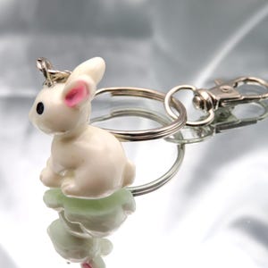 May include: A white rabbit keychain with pink ears and black eyes. The keychain is attached to a silver key ring with a silver lobster clasp.