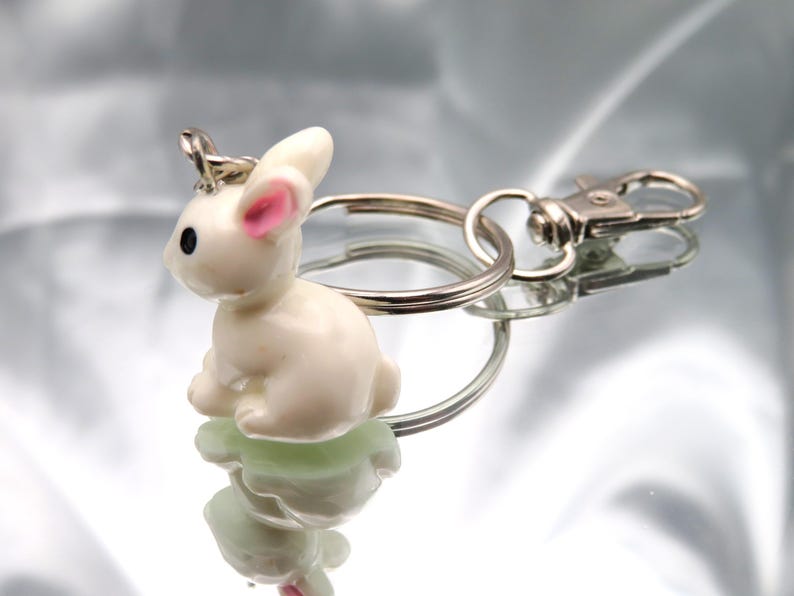 May include: A white rabbit keychain with pink ears and black eyes. The keychain is attached to a silver key ring with a silver lobster clasp.