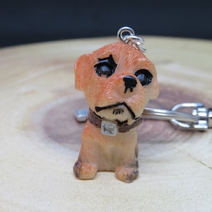 May include: A small, orange-brown, dog-shaped keychain with black eyes and a brown collar. The keychain is attached to a silver metal ring.