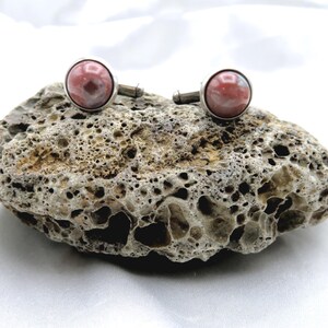 May include: A pair of cufflinks with round, pink and white stone cabochons set in silver-toned metal. The cufflinks are displayed on a textured, gray and brown rock against a white background. The cufflinks are a classic accessory.