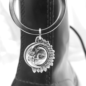 May include: A silver charm featuring a sun and moon design, hanging from a silver ring. The charm is attached to a black boot. The sun and moon are facing each other, with a detailed face on the moon.