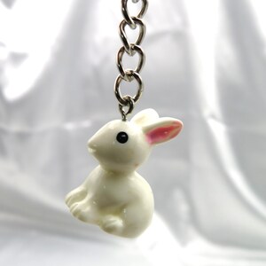 May include: A white rabbit keychain with pink ears and black eyes. The keychain is attached to a silver chain.