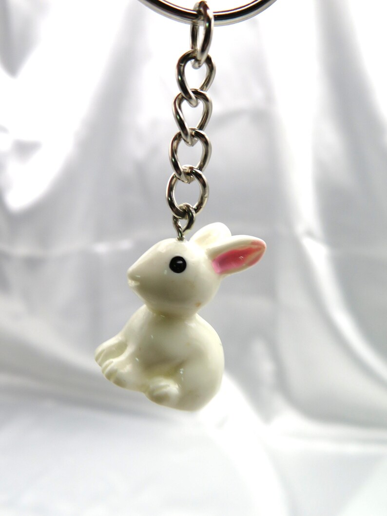 May include: A white rabbit keychain with pink ears and black eyes. The keychain is attached to a silver chain.