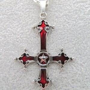 Puede incluir: Collar con colgante plateado con diseño de cruz y esmalte rojo. La cruz tiene una estrella en el centro y detalles decorativos. El collar se muestra sobre una superficie gris claro.