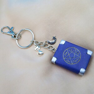 May include: A silver keychain with a blue book charm. The book has a silver border and a pentagram with symbols on the cover. The keychain also has a silver cat charm and a crescent moon charm.