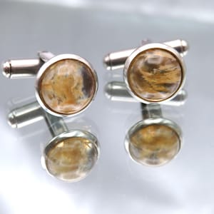 May include: A pair of silver cufflinks with round, marbled, amber-colored centers. The cufflinks are displayed on a reflective surface, showcasing their design and details. The cufflinks are a classic accessory.