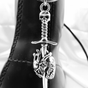 May include: Silver-toned boot charm featuring a skull, a sword, and a heart. The charm is suspended from a metal ring and hangs against a black leather boot. The skull is at the top, followed by a sword piercing a heart.