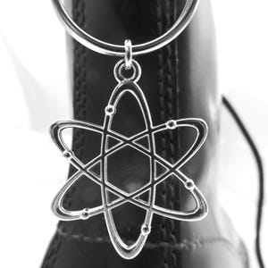 May include: A silver-colored atom charm hangs from a silver ring, attached to a black boot. The charm features a stylized atom design with curved lines and small circular accents. The boot has a glossy finish.
