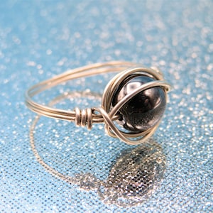 May include: A silver wire wrapped ring with a black gemstone. The ring is on a blue sparkly background.