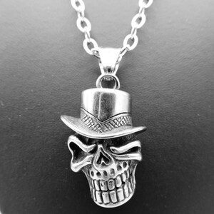 Puede incluir: Un collar con colgante plateado que presenta una calavera con sombrero de copa. La calavera tiene dientes y cuencas de los ojos detalladas. El colgante cuelga de una delicada cadena, creando un accesorio gótico o de inspiración motera.