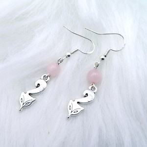 May include: A pair of dangle earrings featuring a silver fox charm and a light pink bead. The earrings have a fish hook style closure. The earrings are displayed on a white, fluffy surface.