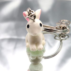 May include: A white rabbit keychain with pink ears and black eyes. The rabbit is sitting on a silver keychain ring.
