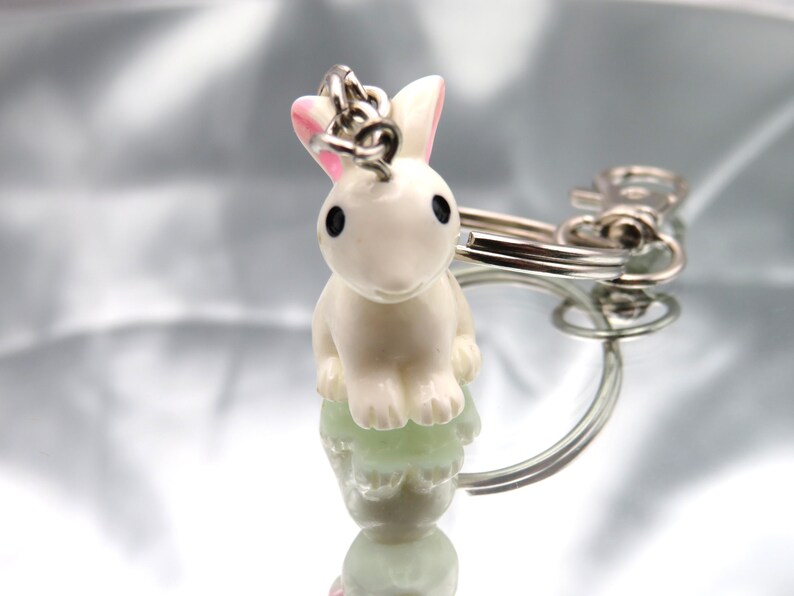 May include: A white rabbit keychain with pink ears and black eyes. The rabbit is sitting on a silver keychain ring.
