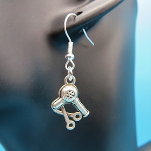 May include: Silver earrings featuring a hair dryer and scissors charm. The earrings are on a mannequin's ear.