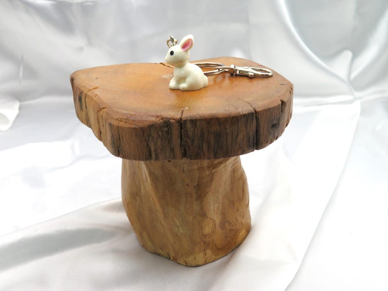 May include: A white ceramic rabbit keychain with a silver metal ring. The keychain is sitting on a wooden stump with a rough, natural surface.