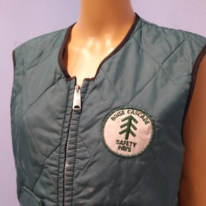 May include: Green quilted vest with a zipper closure and a patch on the front that says "Bouse Cascade Safety Pays" with a green pine tree graphic.