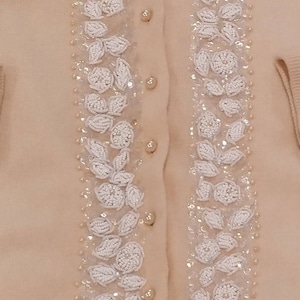Op de afbeelding: Een beige cardigan met witte parelmoer bloemenborduursel aan de voorkant. De cardigan heeft een knoopsluiting.